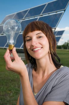 Head and shoulders shot of a young brunette woman,smiling and looking at the camera and holding up a light bulb in front of a free-standing photovoltaic system