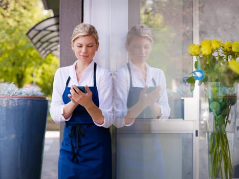Portrait of beautiful caucasian girl self-employed in flower shop, smiling and using mobile phone. Horizontal shape, waist up
