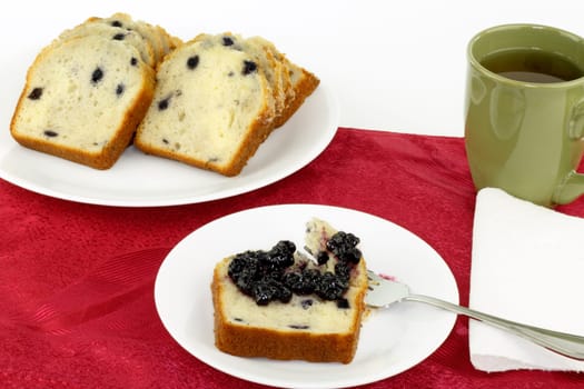 Blueberry preserves spread on a slice of blueberry loaf cake on a white place with a silver fork. A cup of apple juice is on the side as well as the rest of the sliced loaf cake.