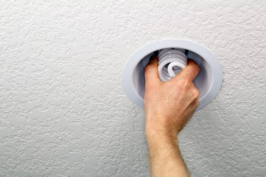 Hand installing spiral compact fluorescent lightbulb in an interior white ceiling fixture.