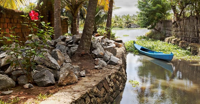 landscape of Kerala backwaters India artery berthed blue canoe local stone granite boulders hibiscus bush with solitory bloom, toe path horizontal with crop space