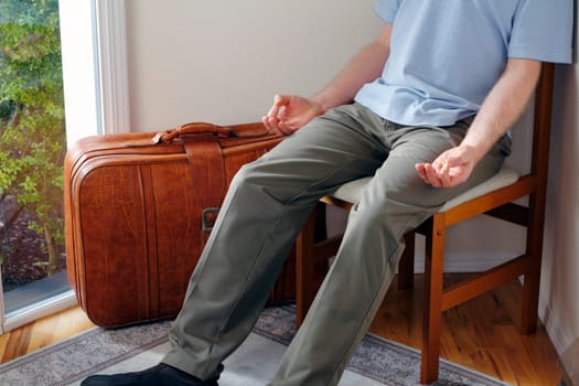 In a home foyer with a suitcase near door a male sits in a chair meditating before traveling to help have a peaceful journey.