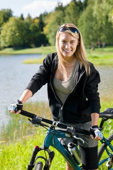 Beautiful young blond woman riding bicycle by lake summer day
