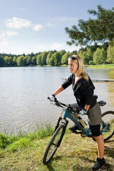 Beautiful young blond woman riding bicycle by lake summer day