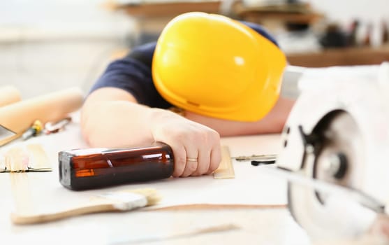 Arm of drunken worker in yellow helmet hold liquor bottle sleeping at table closeup. Manual job workplace DIY inspiration fix shop hard hat industrial education profession career