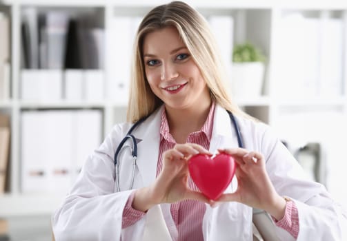 Beautiful smiling blond female doctor hold in arms red toy heart closeup. Cardio therapeutist student education CPR 911 life save physician make cardiac physical pulse rate measure arrhythmia