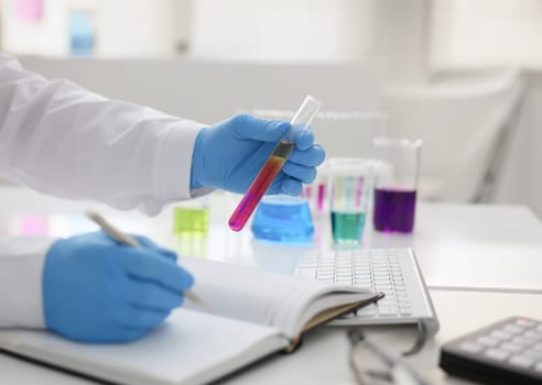 Employee of the chemical laboratory teacher chemist holds a silvery pen in his hand makes notes in the diary records. Test data from reactions examining test tubes with the substance arm in gloves.