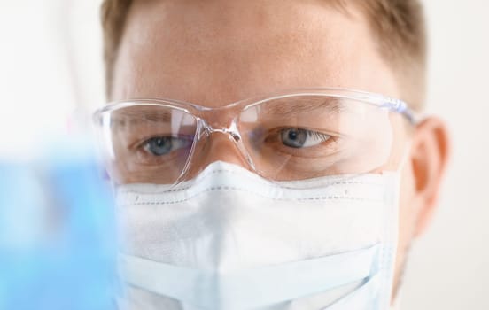 A portrait of a young surgeon chemist doctor looks at a container with a blue liquid and a mask is fought. With viruses and a vaccine for vaccines against diseases.