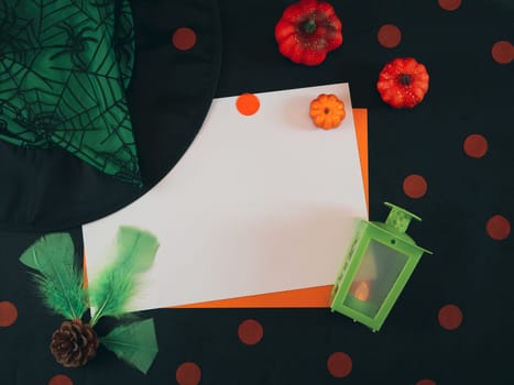 Two empty sheets: white and orange lie horizontally in the middle on a black background with a witch hat, lantern, decorative pumpkins, pine cones, feathers and confetti around. Flat lay close up.