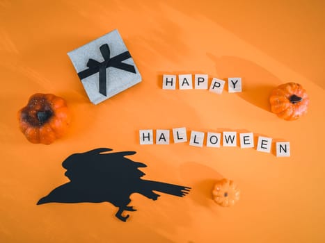 Gift box, three pumpkins, paper witch on a raven and wooden cubes with the inscription: happy halloween on lie on an orange background, close-up flat lay. Concept card greeting.