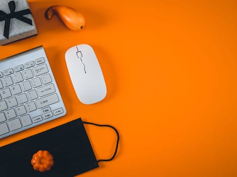 Keyboard, mouse, black medical mask, gift box and pumpkins lie on the left on an orange background with copy space on the right, flat lay close-up. Concept banner halloween.