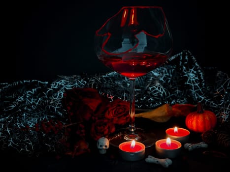 Broken glass with wine, burning candles, skull, bone, dry rose and shiny cape on a black background, close-up side view.Halloween and mystic concept.