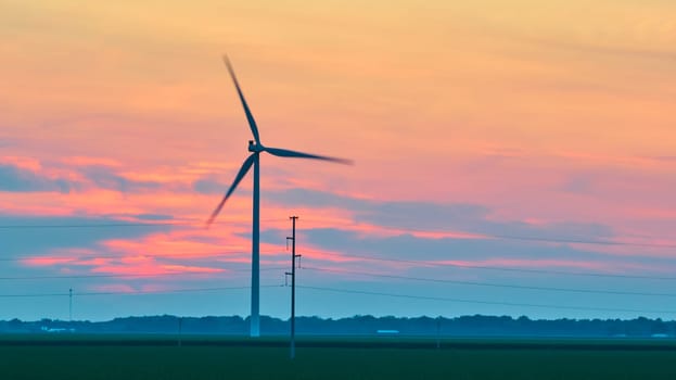 Image of Gorgeous pink and blue sunset clouds behind spinning wind turbine in green field