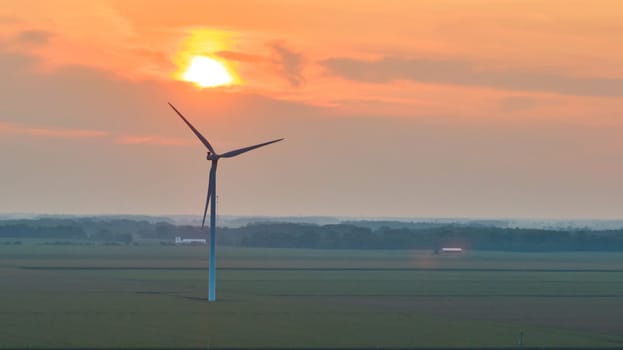 Image of Lone wind turbine in field with orange sun over it at sunset aerial