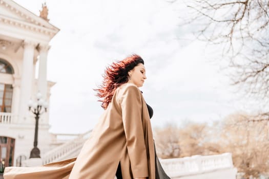 Woman street lifestyle. Image of stylish woman walking through European city on sunny day. Pretty woman with dark flowing hair, dressed in a beige raincoat and black, walks along the building