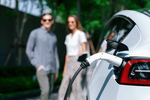 Young couple travel with EV electric car charging in green sustainable city outdoor garden in summer shows urban sustainability lifestyle by green clean rechargeable energy of electric vehicle innards