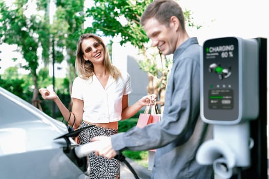 Young couple travel with EV electric car charging in green sustainable city outdoor garden in summer shows urban sustainability lifestyle by green clean rechargeable energy of electric vehicle innards