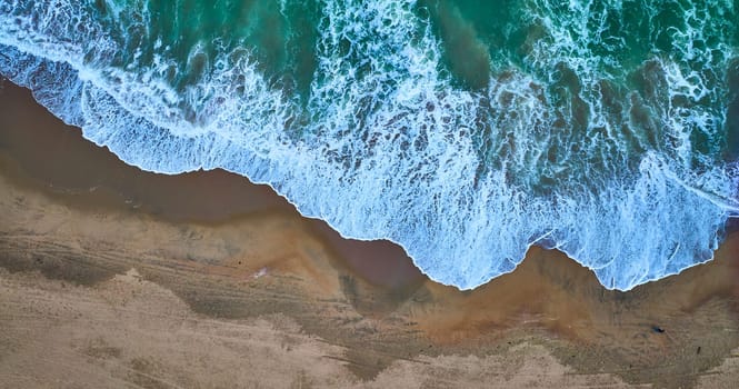 Image of Downward aerial of receding green ocean waves on top with dark tan sandy beach on bottom