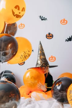 Children's Halloween - a girl in a witch hat and a carnival costume with airy orange and black balloons at home. Ready to celebrate Halloween.