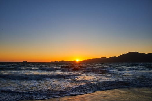 Image of Choppy waves hitting beach and golden sky with sun poking above mountains at sunset