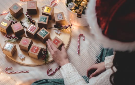 Hands of caucasian teen girl in white knitted sweater takes box of advent calendar with number four while sitting on bed in bedroom, top view close-up with depth of field.