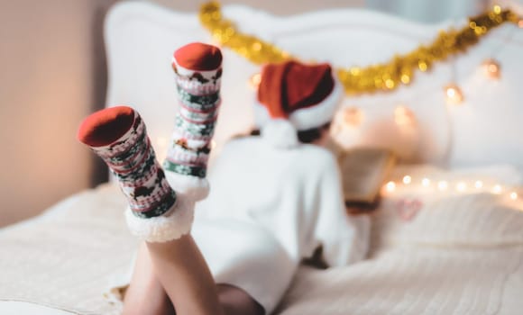 Caucasian teenage girl in a santa claus hat, in a white knitted dress reads a book lying on her stomach, lifting her legs in socks up on the bed in the bedroom, close-up side view with depth of field.