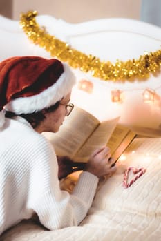 A beautiful caucasian teenage girl in a santa claus hat and white knitted dress reads a book lying on her stomach on the bed against the background of blurry christmas decorations and a garland in the bedroom, side view close-up with depth of field.