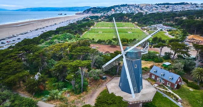 Image of Aerial murphy windmill with small home with soccer fields and city behind it and trees and ocean