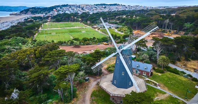 Image of Aerial slate blue murphy windmill with white blades and soccer field behind it with large city