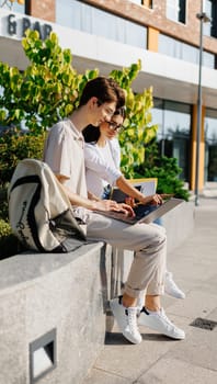 Side view of smiling young students male looking at screen of laptop and female looking down during reading book while sitting together on bench with backpack against blurred green park building