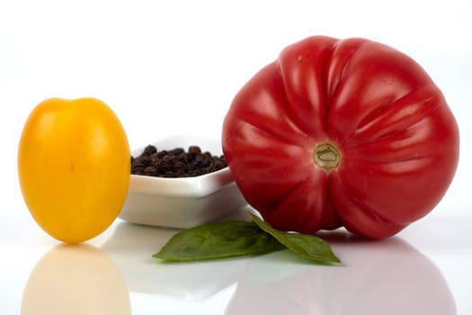 Two different organic tomatoes with small peppercorns skin and basil leaves,close-up mirrored on glass plate. Two different organic tomatoes with a small cup peppercorns and basil leaves on plate glass mirror closeup.