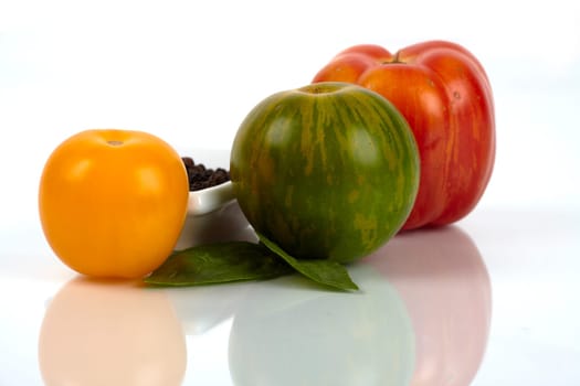 Three different organic tomatoes with small peppercorns,peel and basil leaves,close-up mirrored on glass plate. Three different organic tomatoes with a small cup peppercorns and basil leaves on plate glass mirror closeup.