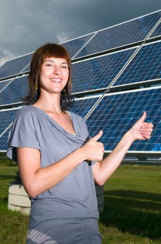 Upper body shot of a young brunette woman in casual casual dress in front of photovoltaic system on green meadow in summer showing thumbs up