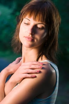 Upper body portrait of a young dark-haired woman in a sleeveless top and crossed arms in front of her chest and hands on her shoulders and lowered gaze on a sidewalk in the park at dusk