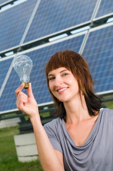 Head-and-shoulder shot of a young woman with a light bulb in her hand in front of a photovoltaic system, looking friendly at the camera.