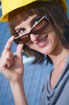 Close-up of a young brunette woman with a yellow construction helmet looking over her sunglasses held with her right hand towards the camera  Background: Solar Panel