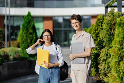 Smiling young male with laptop and female with books looking at camera while standing together on campus street near green trees against blurred college building in daylight