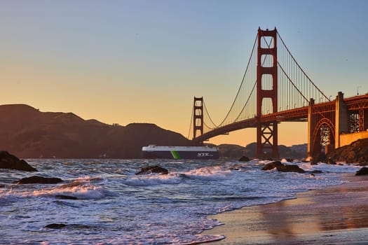 Image of Ocean liner going under Golden Gate Bridge at sunset from sandy beach with waves