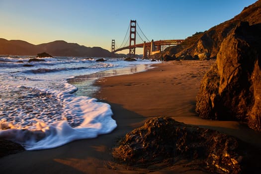 Image of Gorgeous sunset striking Golden Gate Bridge from sandy beach with sea foam on shore