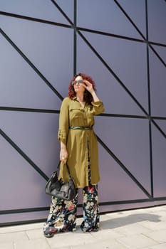 Portrait of a woman standing near a supermarket building. Caucasian model with long brunette hair, wears sunglasses and a khaki dress