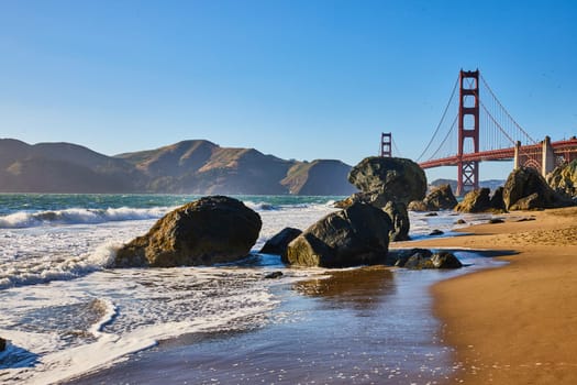 Image of Boulders on sandy beach with waves and seafoam along shore to Golden Gate Bridge