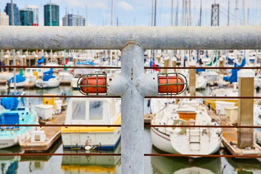 Image of White metal railing with brown iron wires and big heavy duty red lights with blurry boats and city