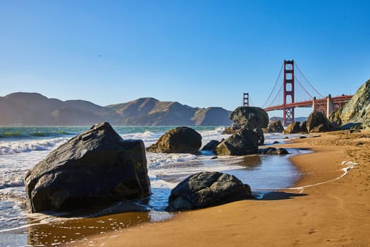 Image of Sandy beach with waves crashing against boulders and distant Golden Gate Bridge