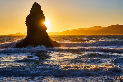 Image of Tall massive boulder jutting out of ocean waves with golden sun poking out from behind