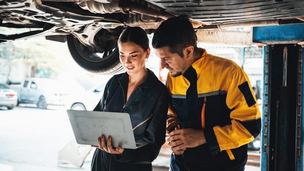 Two vehicle mechanic working together underneath lifted car, conduct car inspection with laptop. Automotive service technician in uniform carefully make diagnostic troubleshooting. Panorama Oxus