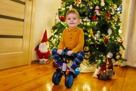 Happy little child boy sitting on push bicycle on a Christmas tree background. Christmas Eve