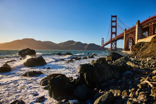 Image of Waves crashing against boulders in shallow water offshore with Golden Gate Bridge at sunset