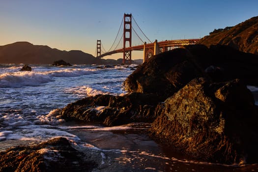 Image of Golden light striking dark mossy rocks with breaking wave and distant Golden Gate Bridge