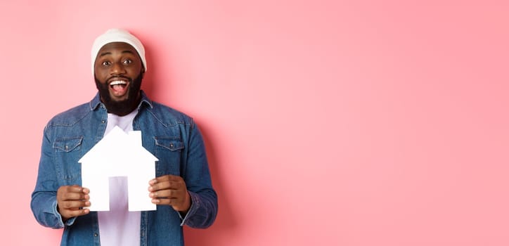 Real estate concept. Happy smiling african-american man holding house model, looking excited at camera, searching for home, standing over pink background.