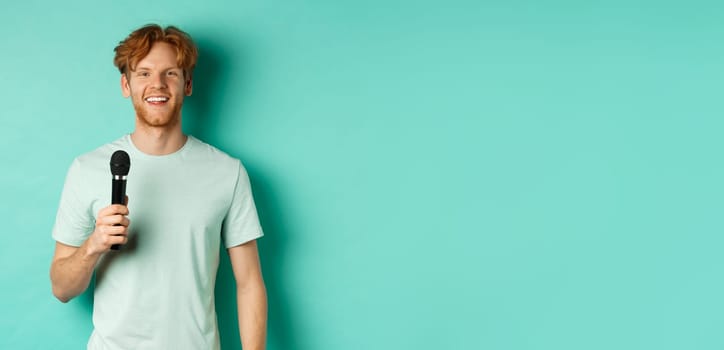 Young redhead man with beard, wearing t-shirt, holding microphone and making speech, singing karaoke, standing over mint background.